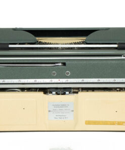 Top-down view of a vintage gray-green typewriter resting on a beige carrying case, with a metal ruler across the front edge and visible typebars behind the carriage.